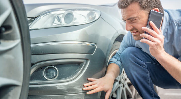 Kemper auto insurance: man talking on the phone while looking at the damage on his car