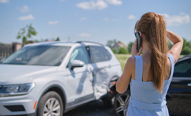 Young woman is on the phone as she looks at her damaged car.