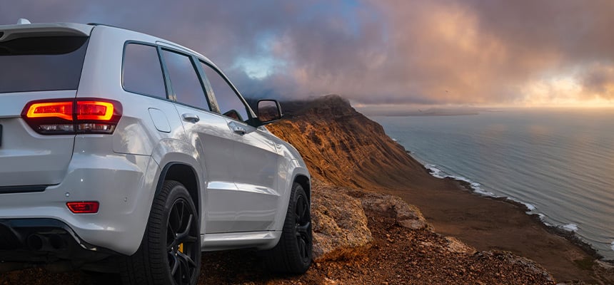 Panoramic, sunset view of a grey SUV on a cliff, facing the ocean